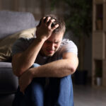 Sad man lamenting sitting on the floor in the living room in a house indoor with a dark background