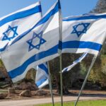 Multiple Israeli flags wave in the wind at a kibbutz, surrounded by lush greenery and historic olive trees. This serene park scene embodies the spirit and traditions of Israeli settlements.