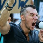 Angry protester using megaphone and shouting while protesting with crowd of people on city streets.