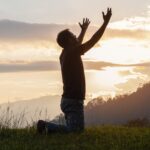 Young male kneeling down with hands open palm up praying to God on the mountain sunset background.
